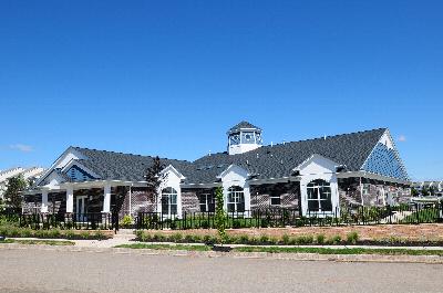 Exterior image of the Harbortown Clubhouse showing a large brick building with a covered entrance. There is a black metal fence surrounding the building, and many bushes, plants, flowers and trees landscape the outside.