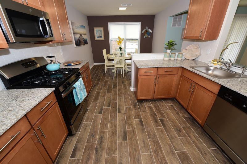 Interior photo of a Harbortown Breeze kitchen showing wood grain tile flooring. Maple color cabinets, granite countertops and stainless steel appliances. There is a table for 4 in the eat in kitchen area.