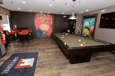 Hawthorne Square Clubhouse photo showing a pool table and a table with four chairs. There is a T.V. on the wall and a Pinball machine in the corner. There are pictures on the wall for decoration.