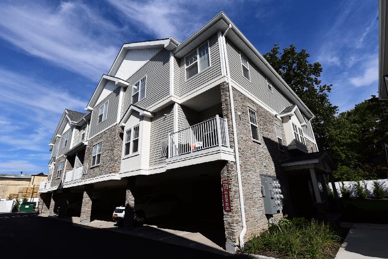 Hawthorne Square Exterior photo showing a brick and vinyl exterior with several balconies. There is parking underneath the apartments. Several bushes and trees highlight the landscape.