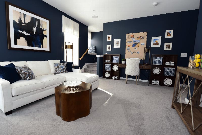 Hawthorne Square Living Room photo showing a couch, center table, a desk and a chair. There are shelves with decorations around the room. There are pictures on the wall above the desk. There is a small staircase leading into the upstairs.