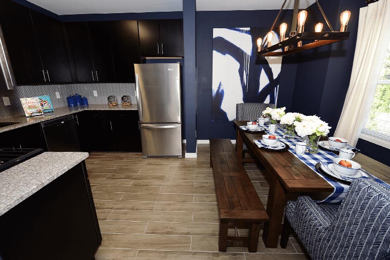 Hawthorne Square Dining Area photo showing the kitchen and dining room. The kitchen has espresso cabinets, stainless steel appliances, granite counter tops and laminate flooring. The dining room has a long table with two chairs and two benches. There are decorations on the kitchen counter and dining room table.