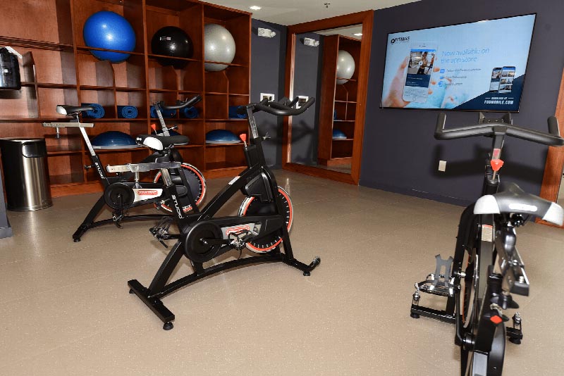 Hawthorne Square Gym photo showing three exercise bikes. There is a T.V. on the wall. There are large shelves on the wall with medicine balls and yoga mats.