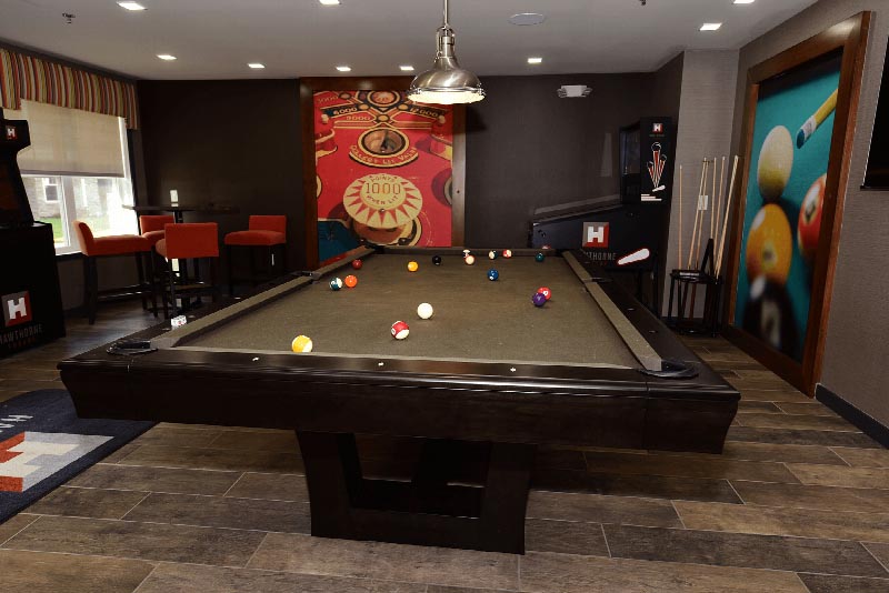 Hawthorne Square Clubhouse photo showing a pool table and a table with four chairs. There is a T.V. on the wall and a Pinball machine in the corner. There are pictures on the wall for decoration.
