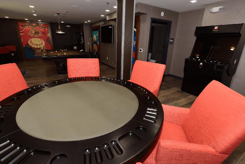 Hawthorne Square Clubhouse photo showing a pool table and a table with four chairs. There is a T.V. on the wall and a Pinball machine in the corner. There is a poker table with several chairs and an arcade game against the wall.There are pictures on the wall for decoration. A bathroom can be seen in the background.