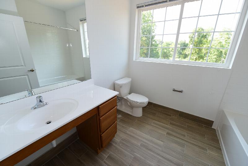 The Grove at Montgomery Bathroom photo with maple color drawers, a white sink and a large mirror on the wall. There is a white toilet and white shower/tub combo and laminate flooring. There is a large window to allow natural light to enter the room.