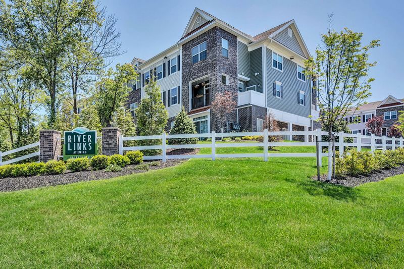 River Links exterior photo showing the River Links entracne sign, with several apartment buildings behind it. There is a grass field, a white wooden fence and lush landscaping