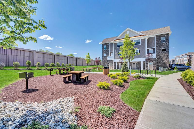 River Links exterior photo showing one of the properties outdoor grilling areas. There are several grills and picnic tables on a stone patio for residents use. There is also a bike rack and several garbage cans.