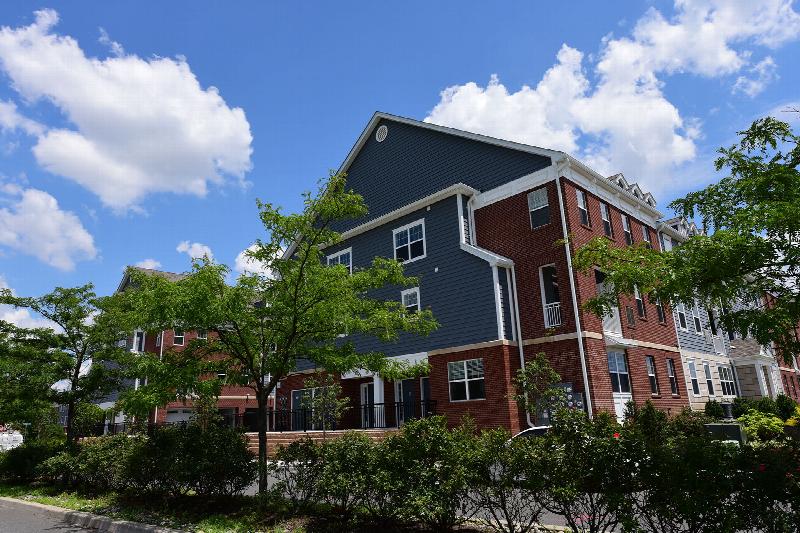 Bayonne Bay exterior photo showing a three story apartment building of red bricks, slate blue colored siding and white trim. Center of photo shows raised walkway with railing leading to several apartment front doors. Street lined bushes and trees highlight an interior street within the community.