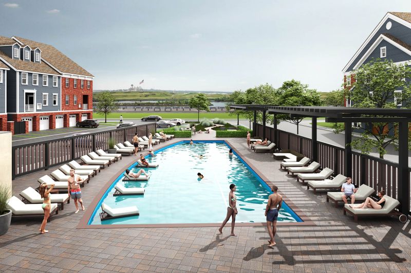Bayonne Bay exterior photo shows residents enjoying the outdoor lap pool with large walk-in sun deck. Spacious outdoor pool area includes lounge chairs and pergola. Displayed in the background are three story townhome apartments with covered balconies. Also shown are garages and parking in front of each unit. Green grass, bushes and trees highlight the surrounding area.