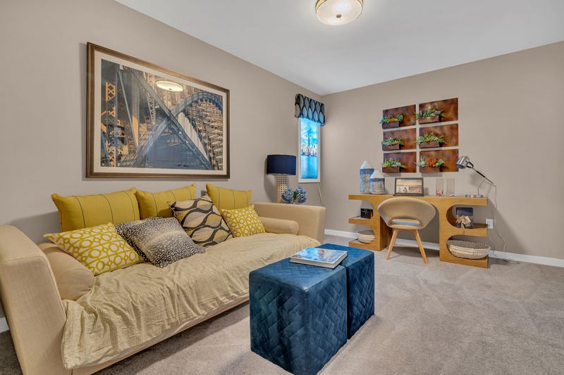 Interior photo of a model apartment at Bayonne Bay showing a den with a large couch with pillows, lush wall to wall carpeting, a window for natural light and a desk.