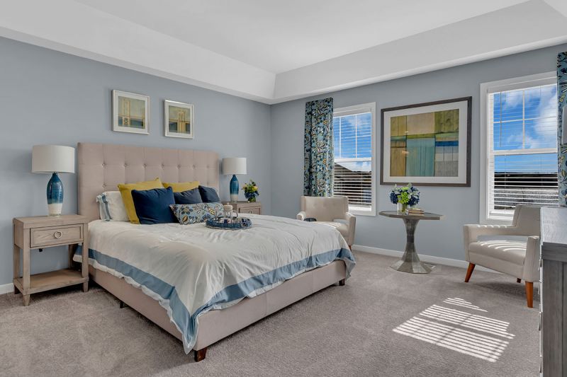 Interior photo of a model apartment at Bayonne Bay showing the primary bedroom with plush wall to wall carpeting, two windows for natural light, a queen sized bed, two nightstands, and a small table with two chairs.