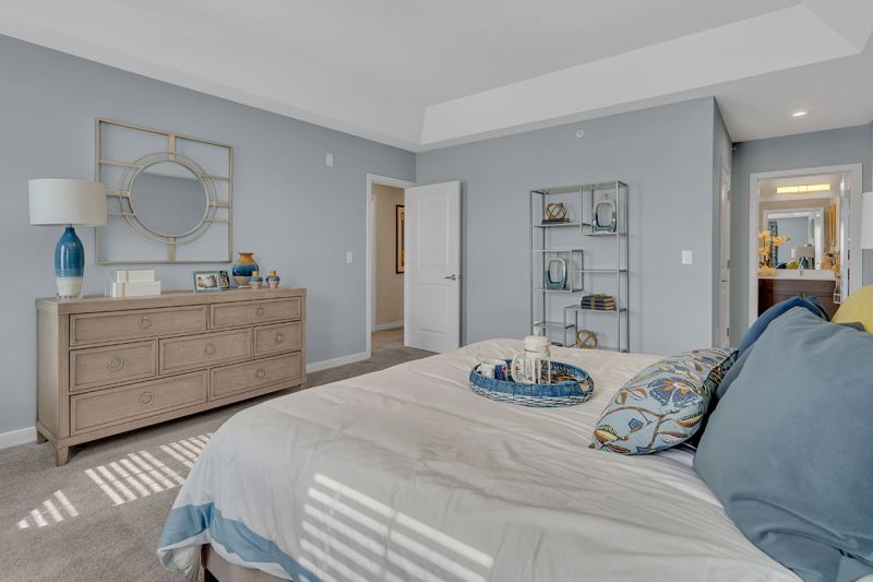 Interior photo of a model apartment at Bayonne Bay showing the primary bedroom with plush wall to wall carpeting, queen sized bed, large dresser and an entry into the primary bathroom.