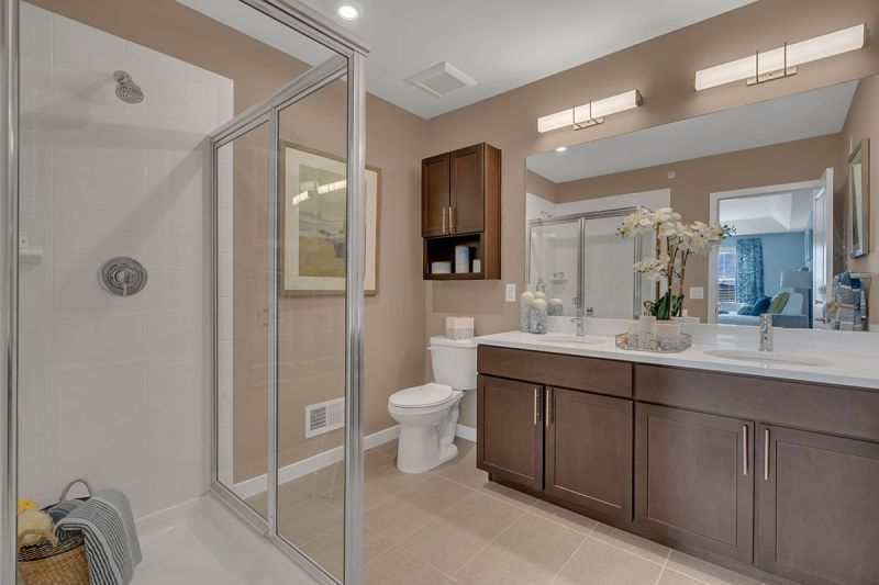 Interior photo of a model apartment at Bayonne Bay showing the primary bathroom with ceramic tile, dual sinks with vanity, large mirror, stand up shower and toilet with medicine cabinet above.