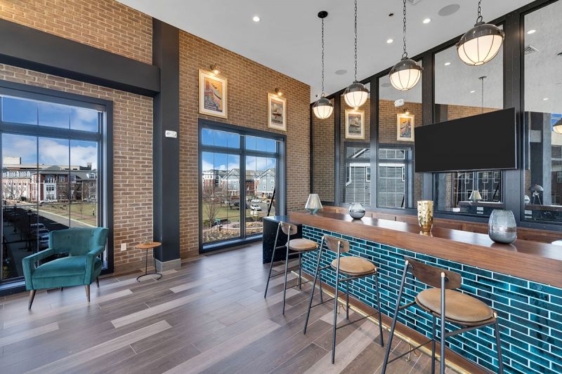 Interior photo of The Club at Bayonne Bay showing the Party Room, featuring luxury vinyl plank flooring, and a bar with stools and a TV hung behind it, as well as large dramatic windows overlooking the community.