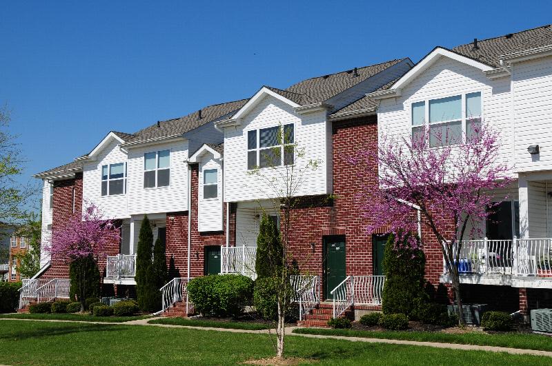 Exterior image of Harbortown Ports North East showing a red brick apartment building with white vinyl siding and trim. There are multiple staircases leading to entrances with green doors. Bushes, flowering trees and green grass landscape the area.