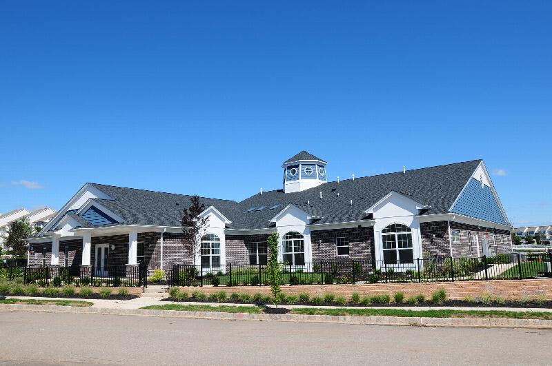 Exterior image of the Harbortown Clubhouse showing a large brick building with a covered entrance. There is a black metal fence surrounding the building, and many bushes, plants, flowers and trees landscape the outside.