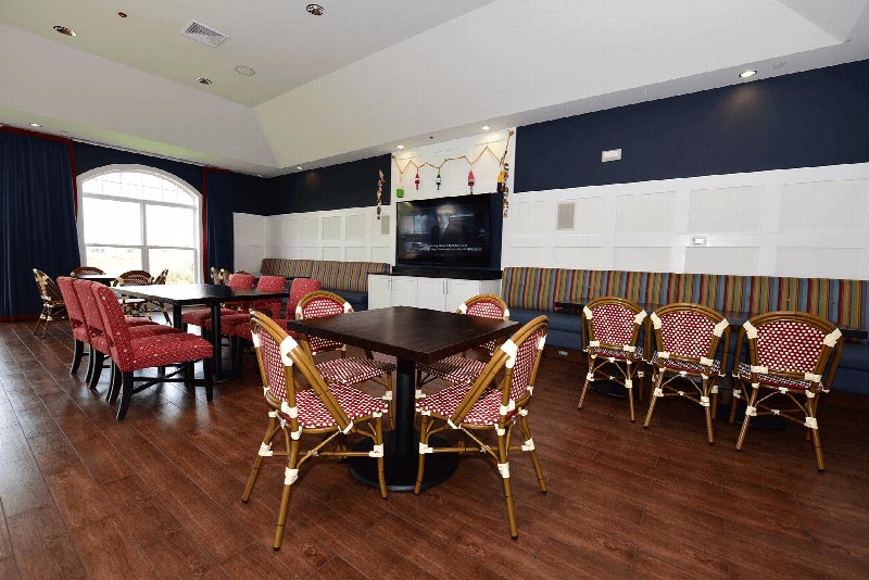 Harbortown Clubhouse community room photo showing multiple seating areas and a large screen TV mounted on the wall. There is hardwood floors and a large window to allow for natural light.