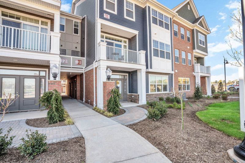 Exterior photo of a typical 3 story building at Ewing Town Center showing the exterior finishes, including red brick, mixed stone and blue siding with white windows and trim. Brick pavers lead from the sidewalk to the entry doors. Lush landscaping surrounds the building.