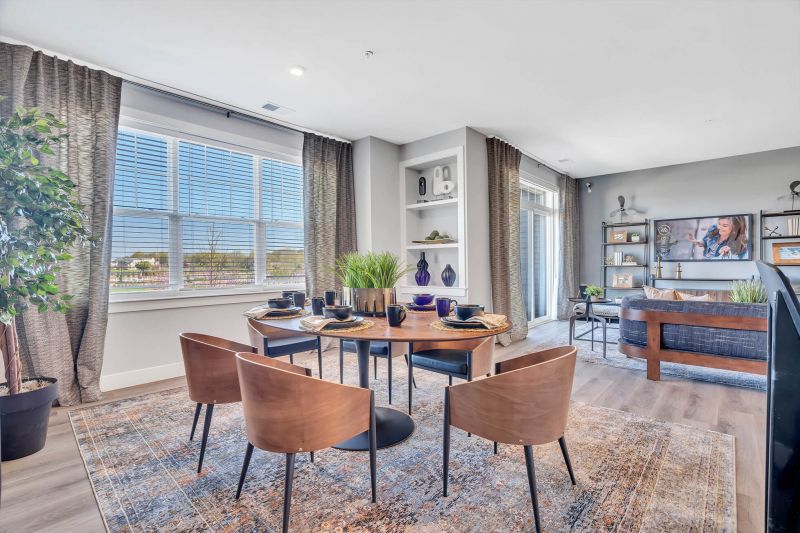 Interior photo of the dining room area in a tyical apartment at Ewing Town Center showing a table set for six, with a large window allowing in natural light. The living room is shown in the background.