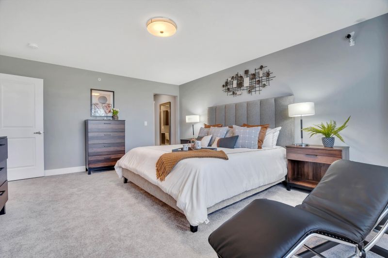 Interior photo of the Primary Bedroom suite in a typical apartment at Ewing Town Center showing a bed, several dressers, end table with lamp, a lounge chair. There is a doorway showing access to the primary bath