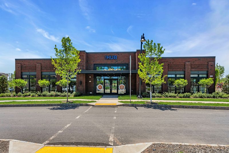 Exterior photo of the clubhouse at Ewing Town Center, showing a large brick building with black windows and trim, and a wide pathway leading to a covered double door entry.