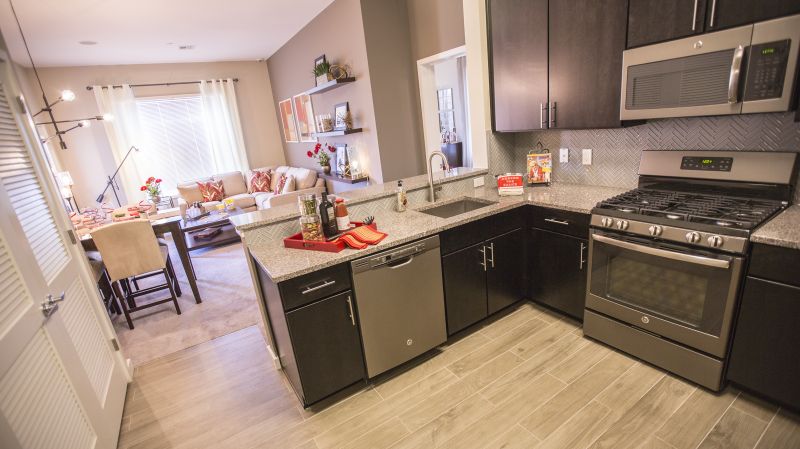 Grove Crossing living room photo showing the living room from the kitchen. The kitchen has espresso cabinets, stainless steep appliances and laminate flooring. There is a sectional couch and small table in the living room. There is a 4 person dining room table and several chairs. There are decorations on the kitchen counter and the tables.
