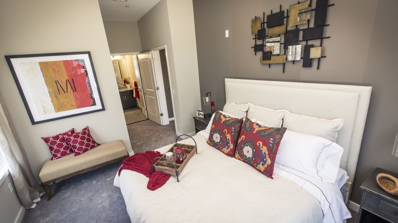 Grove Crossing bedroom showing a queen sized bed, two nightstands with lamps, and a bench against the wall. There are decorations on the nightstands and the bed. The bathroom can be seen in the background.
