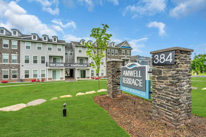 Exterior photo of Amwell Terrace showing a 3 level apartment building with gray siding, mixed stone and white trim. There is a large lawn and the monument sign for Amwell Terrace can be seen in the foreground.