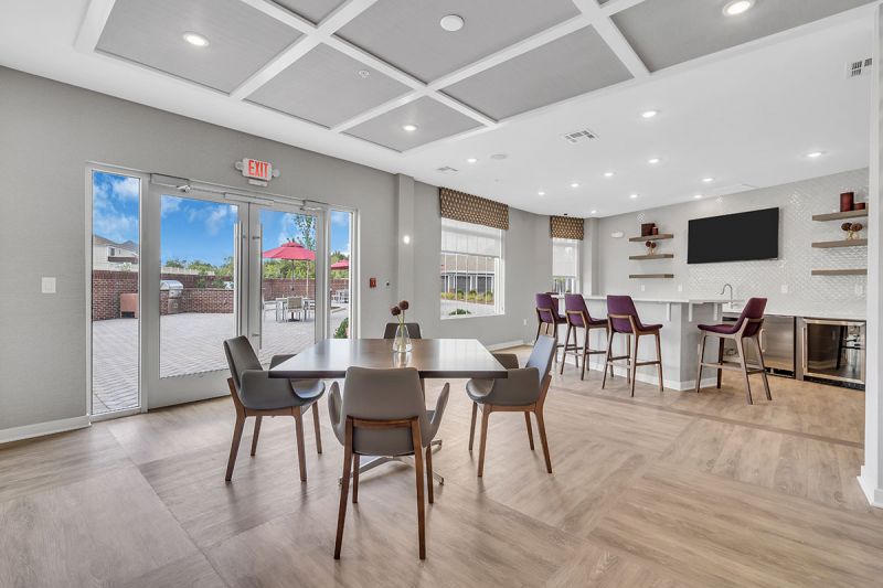 Interior photo of one of the community spaces at Meadows at Montgomery showing a luxury vinyl tile floor and multiple seating areas. Ther is a large screen TV on the wall, and doors leading out to the community patio with seating and grills.