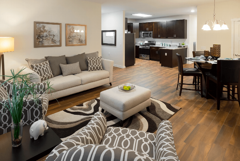 Gardens at Jackson Twenty Interior photo showing the living room, dining room and kitchen. There is a couch, a chair and an ottoman in the living room. There is a 4 person table and chairs in the dining room. The kitchen shows espresso cabinets and stainless steel appliances. Laminate flooring runs the entire apartment.