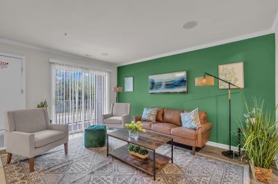 Interior photo of the living room at Meridian Terrace showing modern white oak flooring, a couch and two chairs with a coffee table. A glass sliding door with vertical window blinds leading to the balcony overlooking the community’s exterior.