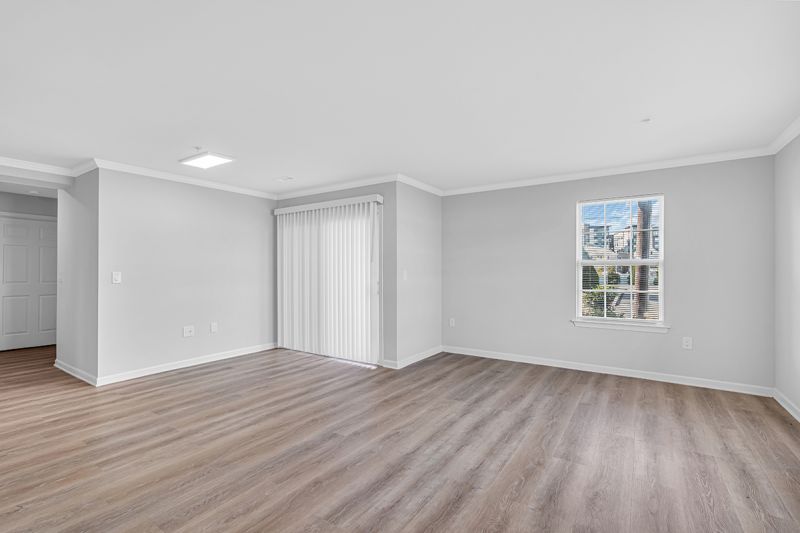 Interior photo of the living room at Meridian Terrace showing modern white oak flooring with large windows allowing natural light throughout the entire unit. A glass sliding door with vertical window blinds leading to the balcony can also be seen.