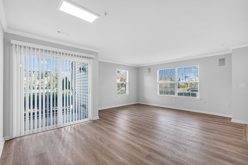Interior photo of the living room at Meridian Terrace showing freshly painted walls, modern white oak flooring with large windows allowing natural light throughout the entire unit. A glass sliding door with vertical window blinds leading to the balcony can also be seen.