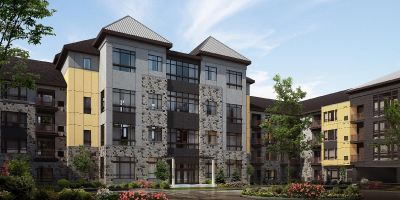 Next at Xchange exterior photo shows five story apartment building with light colored brick and stone facade. Garden style shrubs, green grass and trees highlight the surrounding landscaping