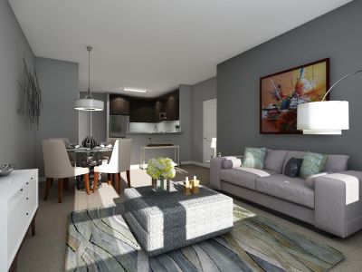 Next at Xchange interior photo of living room shows a couch and square ottoman facing a TV console. Adjacent to the living room is a dining table that seats up to four. Both the living and dining room area is decorated with artwork and complementary accessories, it is carpeted with the exception of the kitchen area seen in the background. The kitchen has expresso cabinets, stainless steel appliances and wood like floor tiling.