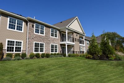 Exterior photo of the exterior of an apartment building in the affordable housing section at The Club at Woodhaven, showing a 2 story building with tan siding, white trim and windows and mixed stone. Lush landscaping surrounds the area.