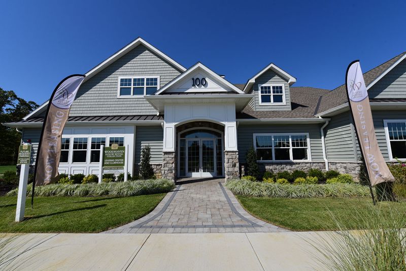 Exterior photo of the clubhouse at Woodhaven, showing green siding, stone walls, white trim and a covered front entrance. The areas is lushly landscaped and accessed by a paver walkway.