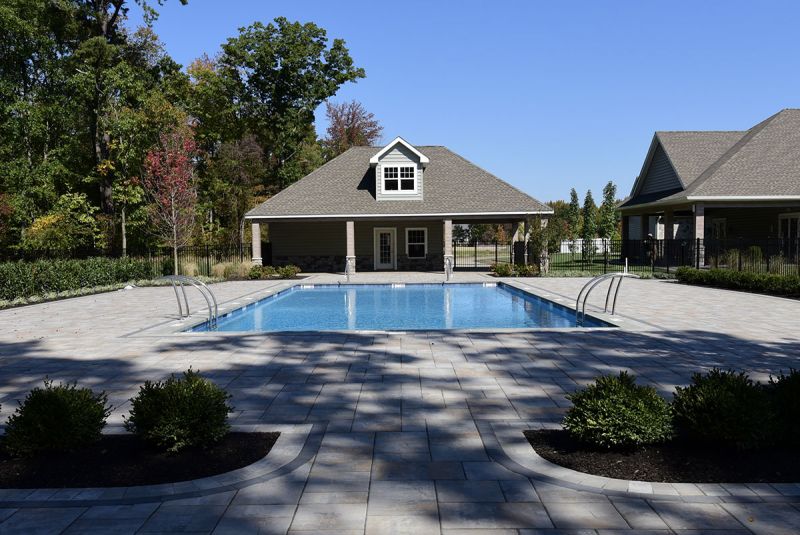 Exterior photo of the pool area at the clubhouse at Woodhaven showing a large swimming pool surrounded by a paver patio and lush landscaping with a large pool house.