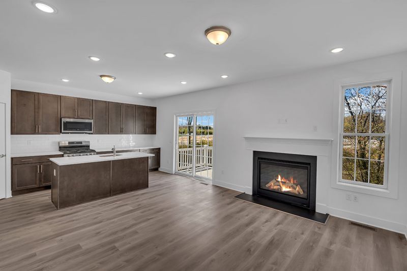 Interior image of a townhome at the Club at Woodhaven showing luxury vinyl woodgrain plank flooring in the gamily room and kitchen area. There is a large gas fireplace and window in the living room and sliding glass doors to an outside deck. The kitchen features new cabinets, stainless steel appliances and an island with sink.