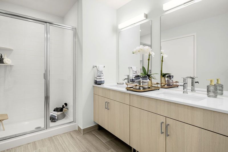Interior photo showing a typical master bathroom in an apartment at RVR with ceramic tile woodgrain flooring, vanity with 2 sinks and mirrors, and a large walk in shower.