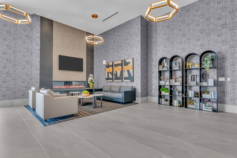 Interior photo of community lounge located on the 5th floor at RVR showing a large couch, coffee tables, and two additional single couches in the middle of the room. To the left is an electric fireplace and above is a mounted TV. To the right are four stand-up shelves with multiple decorative pieces. Multiple overhead light fixtures, artworks, and modern wallpaper can be seen in the vicinity.