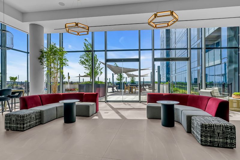 Interior photo of the lounge area on the 5th at RVR floor showing two large sectional couches, small round tables in front, floor-to-ceiling windows, and multiple light fixtures overlooking the community’s exterior.