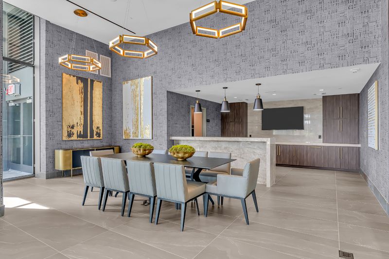 Interior photo of community room located on the 5th floor at RVR showing a professional demonstration kitchen with a large bar-height marble counter, mounted TV, and built-in wooden cabinets. To the left of the demonstration kitchen is a large dining table seating 10 people. Multiple light fixtures, modern wallpaper, and art paintings can be seen in the vicinity.
