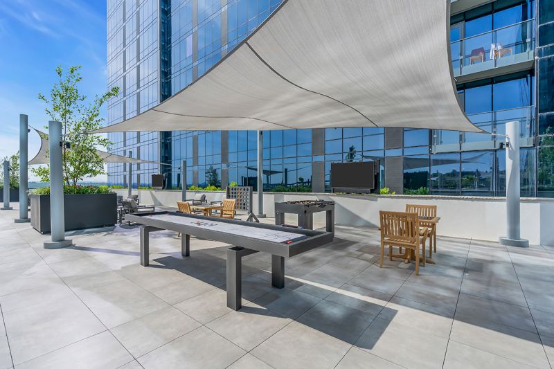Exterior photo of the rooftop located on the 5th floor at RVR showing a shaded gaming area including air hockey table, foosball table, giant connect 4 sets, and built-in chess tables with chairs. The community building can be seen in the background.