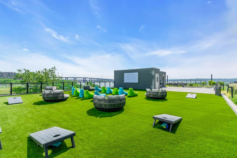 Exterior photo of the rooftop located on the 5th floor at RVR showing an outdoor theater/ gaming area on grass including multiple wooden cornhole sets surrounded by modern patio furniture and bean bag chairs. To the left is a utility room with a large mounted TV. Amazing views can be seen in the background.