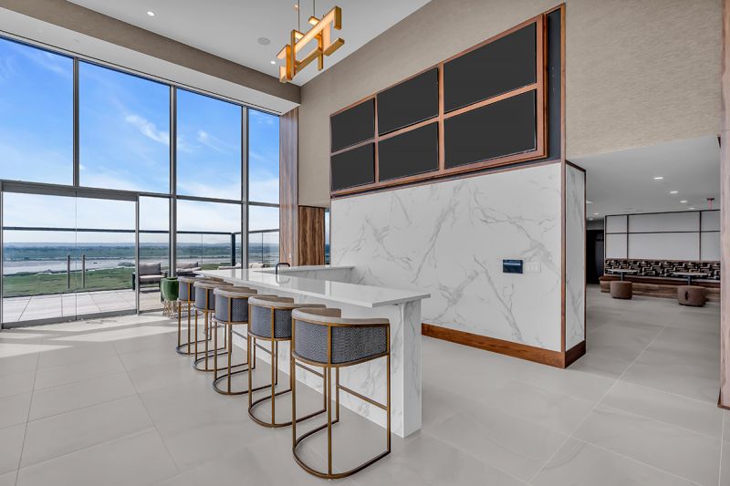 Interior photo of community room located on the 19th floor at RVR showing another angle of marble counter, bar-height seating, and multiple mounted TVs in the center of the room. To the left are clear glass double doors leading to the exterior of the community. Additional seating areas can be seen in the background.