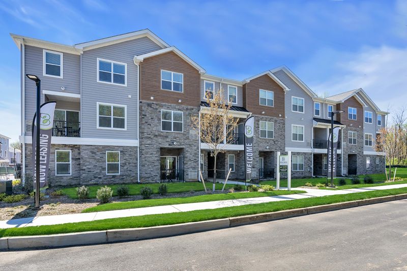 Exterior Photo of The Boulders at Beekman showing a 3 story apartment building with gray and tan siding, mixed gray stone, white trim and black balcony railings. Sidewalks and fresh landscaping surround the building.