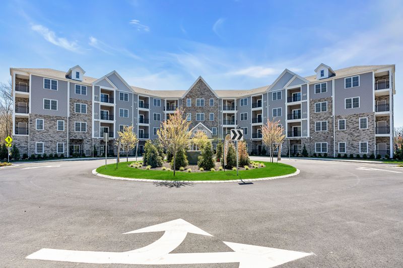 Exterior photo of an apartment building at the Enclave at Woodhaven, showing a 4 story building with gray siding, white trim and mixed stone. Lush landscaping can be seen surrounding the building.