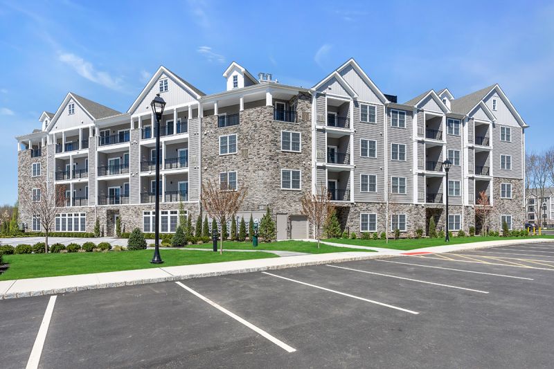 Exterior photo of an apartment building at the Enclave at Woodhaven, showing a 4 story building with gray siding, white trim and mixed stone. Lush landscaping can be seen surrounding the building.